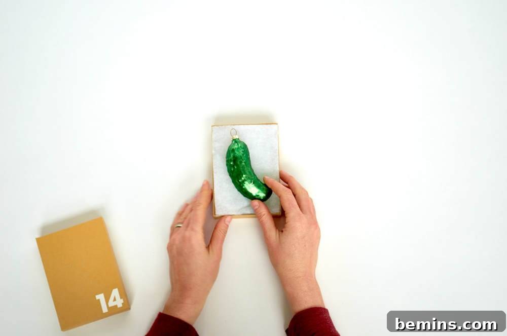 Two hands hold a small white object with a green attachment on it next to a tan box with the number 14 on it.