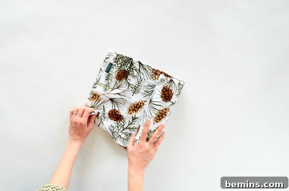 Two Women's Hands Holding a Folded Piece of Cloth Decorated with Brown Pine Cones and Green Tree Twigs, Wrapping a Bottle