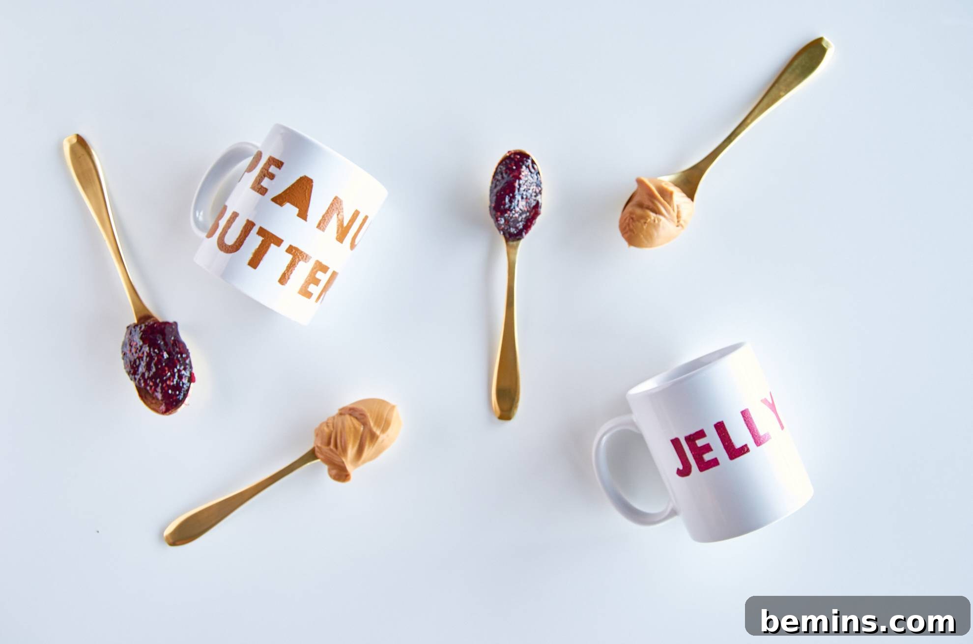 Two spoons with jelly and two spoons with peanut butter scattered about a mug that says peanut butter and a mug that says jelly.