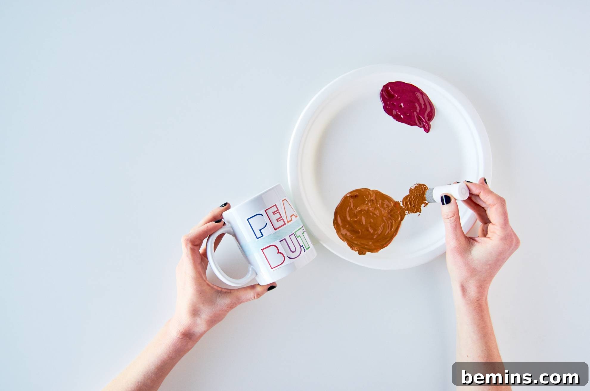 Woman holding a mug in one hand while placing craft paint onto a paper plate with her other hand.