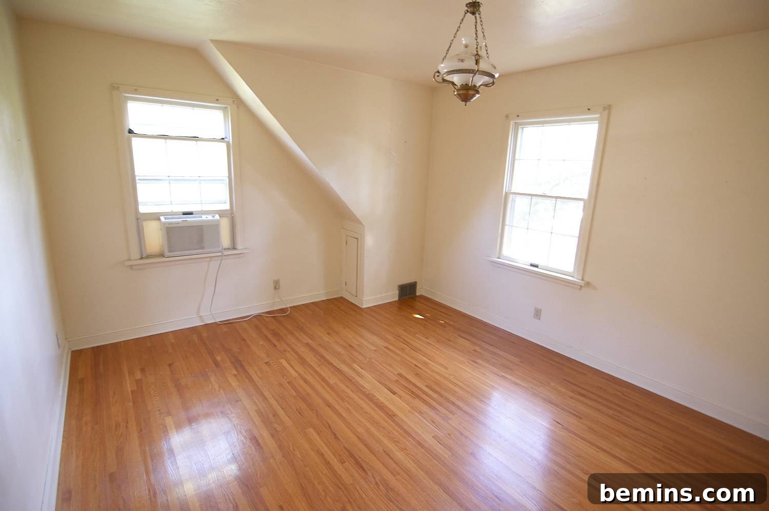 Interior of a room with two windows, white walls, hardwood floors, and a light fixture hanging from the ceiling. Before makeover.