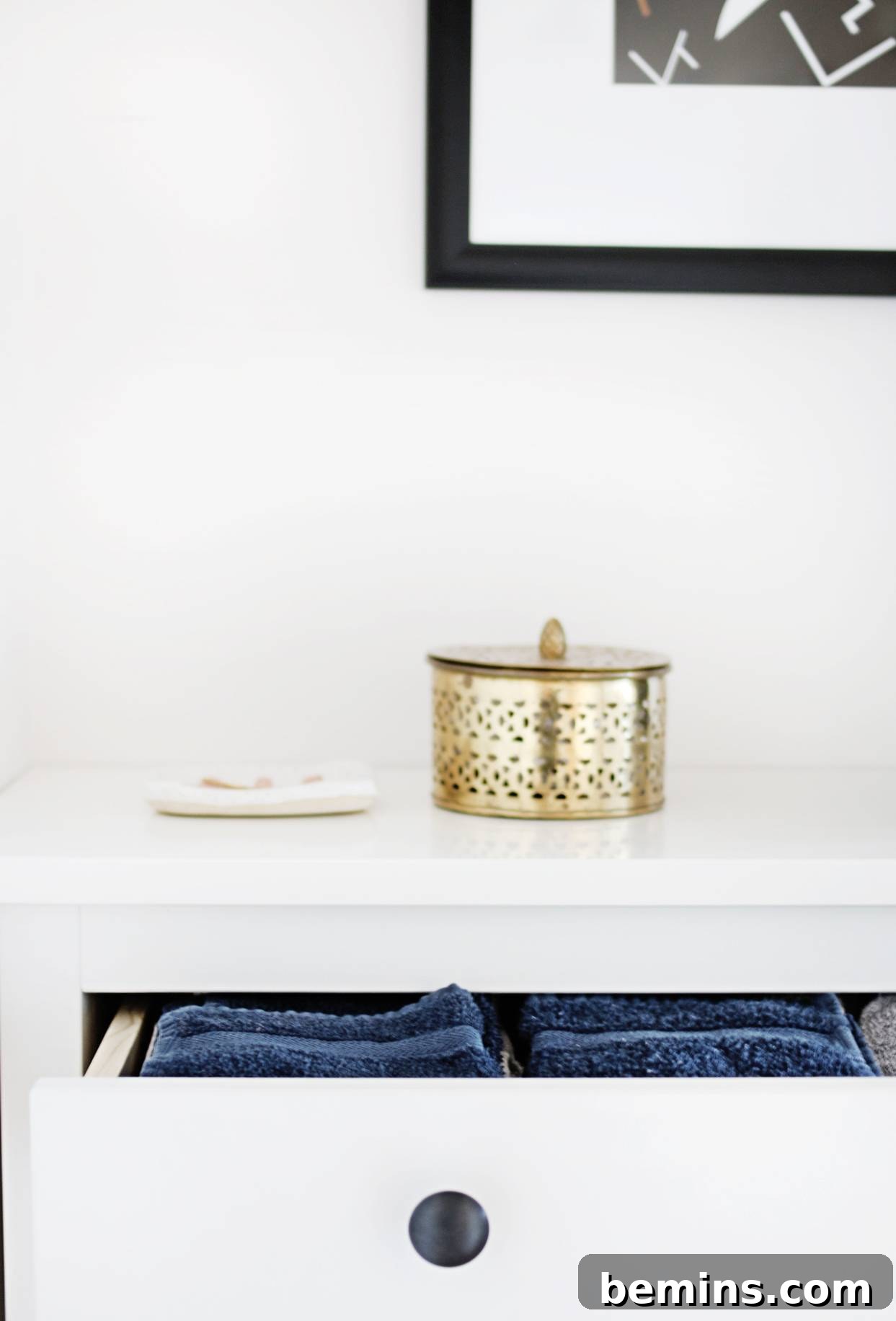 An open top draw of a white dresser with blue towels in it and a gold chatchky on top. The HEMNES shoe cabinet is repurposed for towel storage in the hallway.