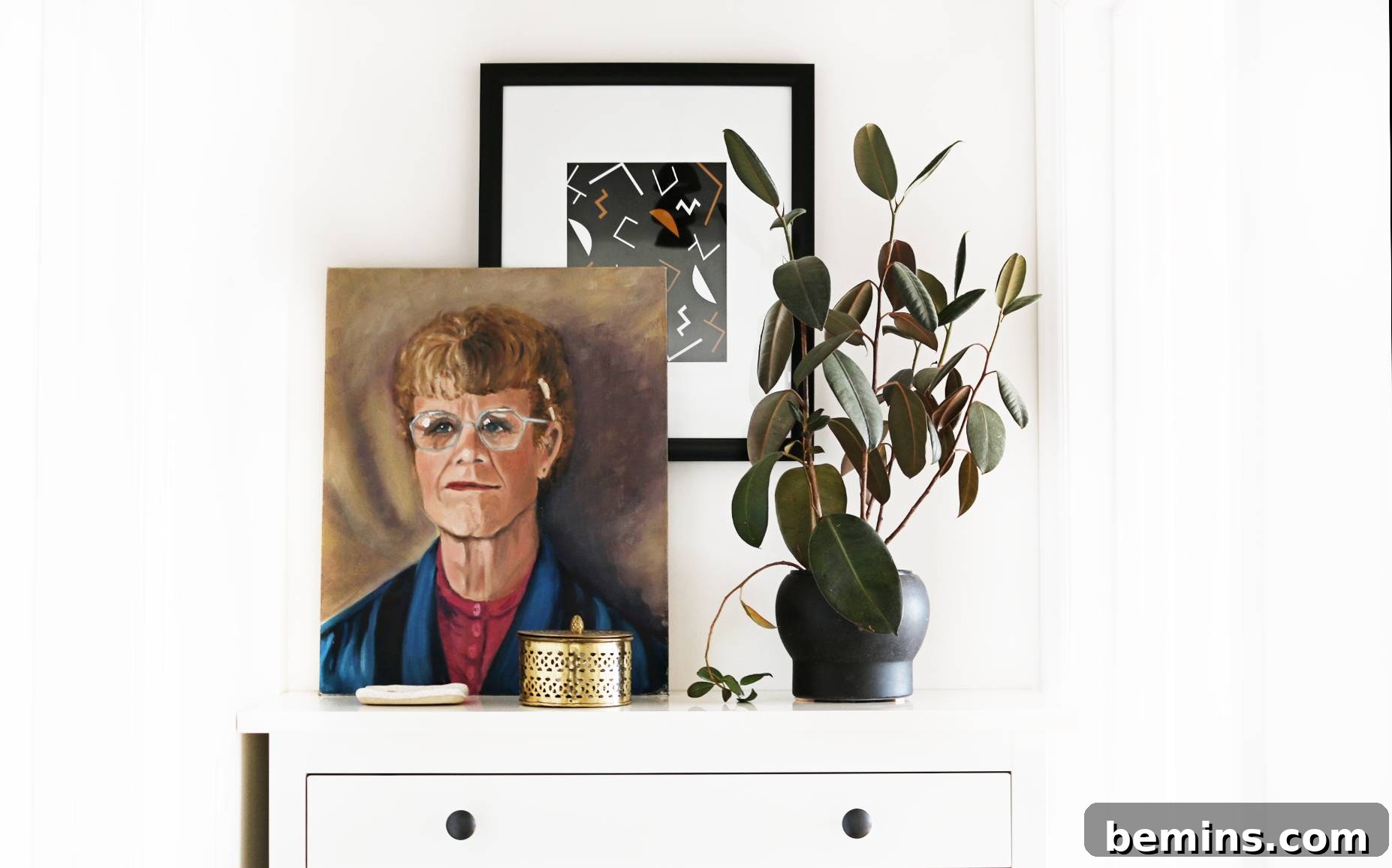 On a white chest of drawers, there is a potted plant on the right and a painting of a person to the left, with another picture visible behind it. Art and plants add personality and charm to the hallway's decor.