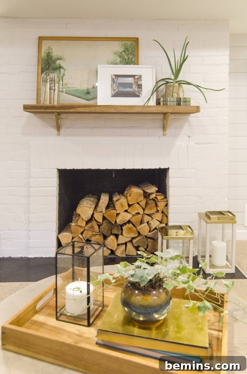 A fireplace stacked full of firewood on a white ship lapped wall, a wooden serving tray with a candle, potted plant and books, more candles by the fireplace.
