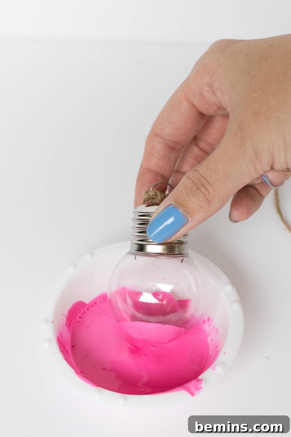 Close-up of a hand with vibrant blue nail polish carefully holding a clear ornament filled with swirling pink liquid paint.