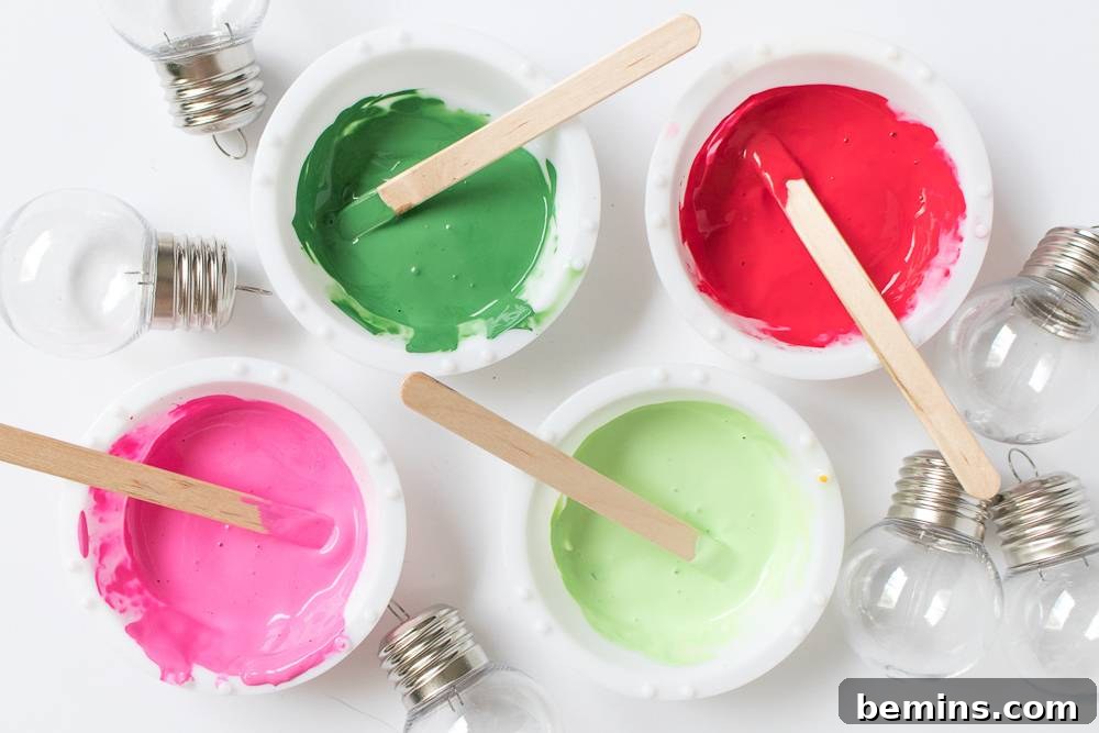 Overhead shot of four small bowls filled with vibrant red, dark green, lime green, and hot pink paints, each with a popsicle stick for stirring, surrounded by soft string lights.