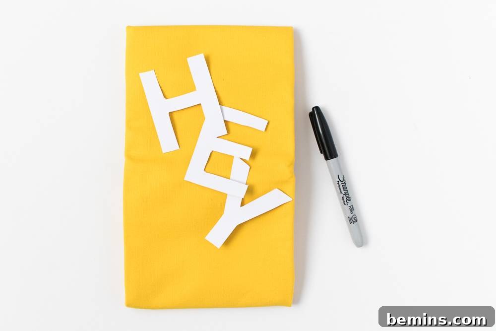 Cut out letters are on a yellow sheet of paper, with a black pen nearby. Preparation for the DIY pillow project.
