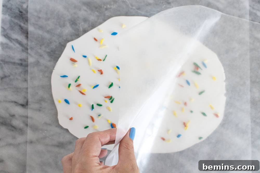 A woman's hand with blue nailpolish is pulling a piece of wax paper over a white rolled out piece of dough with multi colored pieces spread over it.