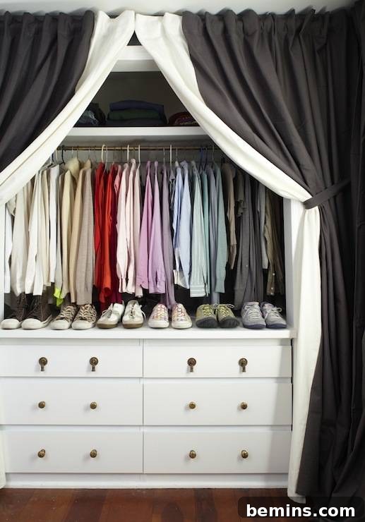A black and white curtain tied back to reveal a neat closet with clothes hung by color and shoes lined atop a white dresser.