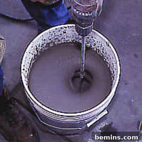Person expertly mixing concrete with a drill and paddle in a bucket.