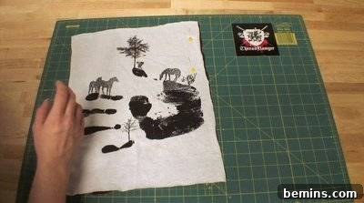 A white piece of cloth with grey horses and a handprint on it on top of a green cutting mat. Sewing T-Shirts.