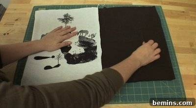 A person's hand holding down a white piece of cloth with a black handprint and horses, and a black piece of cloth, on a green cutting mat. DIY Sewing Project.