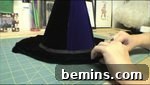A woman works on the brim of a black witches hat that is on a cutting mat, adding bias tape to the edge.