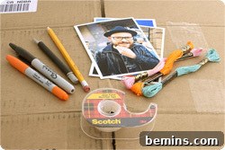 Photographs sit on and near packages of yarn, markers, a roll of tape and a flattened cardboard box.