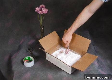 A person's arm going into cardboard box filled with flower next to a roll of wide tape, a pair of scissors and a little vase with tall pink flowers in it.