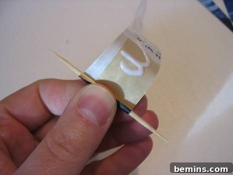 A close-up of hands carefully winding a pre-glued, tapering paper strip around a toothpick, starting from the wide end, illustrating the technique for forming a paper bead. The paper is wrapping tightly and evenly around the toothpick.