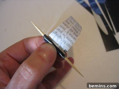 A person's fingers gently holding a toothpick with a partially wound paper strip, showing the progress of rolling a paper bead. The strip is coiling tightly and uniformly around the toothpick, demonstrating proper winding technique.