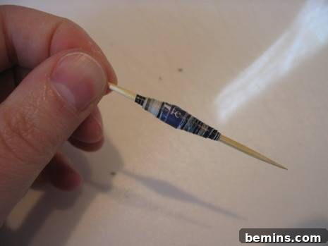 A close-up of a hand gently holding a toothpick with a freshly rolled paper bead, demonstrating the careful process of slipping the bead off the winding tool. The bead is intact and ready for drying.