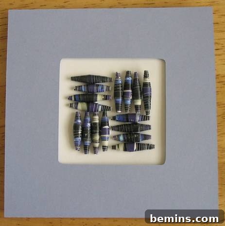A close-up of a meticulously matted square artwork featuring an intricate geometric design composed entirely of tightly rolled, colorful paper beads. The beads create a textured, three-dimensional pattern at the center of the piece.