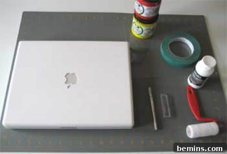A well-organized workspace for DIY stencil printing, featuring a white MacBook displaying a design, rolls of masking tape, vibrant fabric paints, and a small roller on a grey cutting mat, ready for crafting.