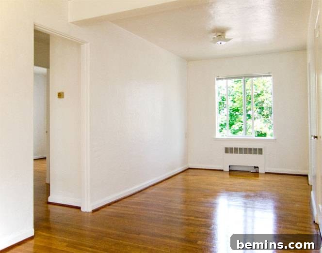 A bright empty room with white walls and glossy wooden floors, and a single window.