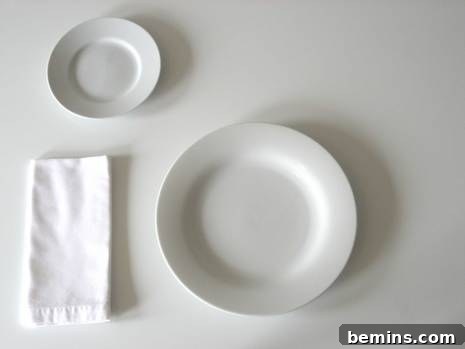 A white bread plate positioned above and to the left of a larger white dinner plate, with a folded white napkin.