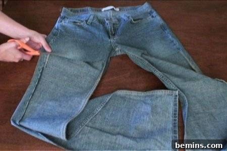 How to Sew Your Own Overalls 25 Woman cutting a pair of blue jeans on top of a wooden table using orange handle scissors.