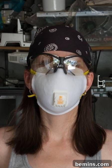 A woman in a gray tank top wears a white mask covering her nose, mouth and cheeks, as well as safety glasses and a black bandana on her head. Staying safe while doing DIY projects.