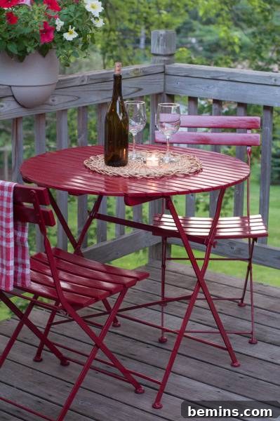 A small round red metal table on a fenced deck, with a wine bottle and two wine glasses on it, and two red chairs around it. A perfect outdoor dining setup with a vibrant red bistro table.