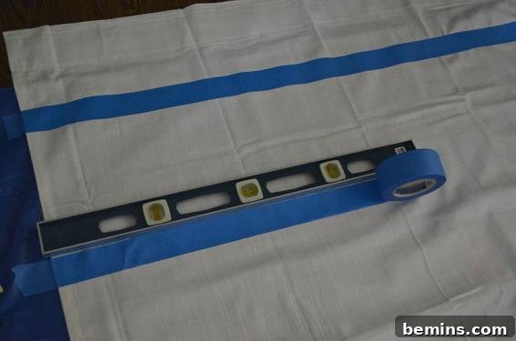 Painted Stripes: A Curtain Refresh 6 A piece of white cloth with a blue stripe, a level laying on the cloth next to a strip of blue tape, illustrating the careful application of tape for crisp lines.