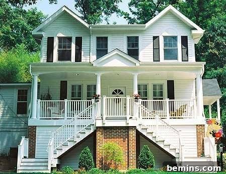 A large, white three-story home nestled among trees, showcasing the successful integration of a second story addition and a welcoming front porch, resulting in a stunning transformation