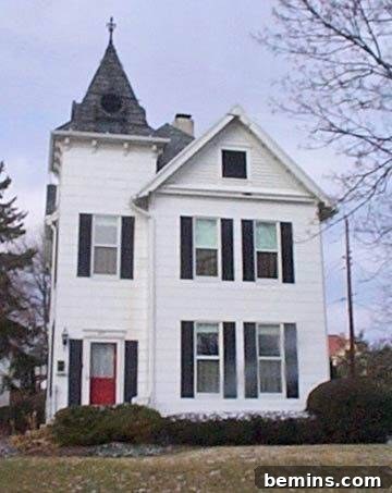 A beautifully restored white two-story Victorian home with a vibrant red door and an ornate rooftop cross, showcasing the charm of a wraparound porch