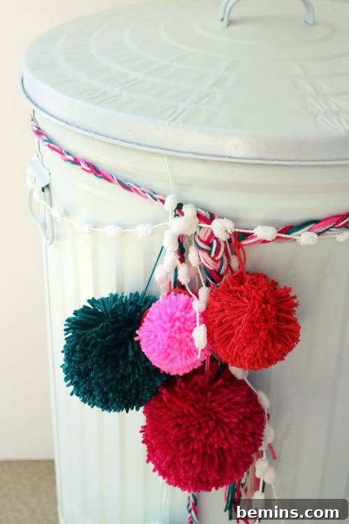 Brighten Up Your Bin: A Trash Can Transformation 3 A garbage can painted in white decorated with pom poms.