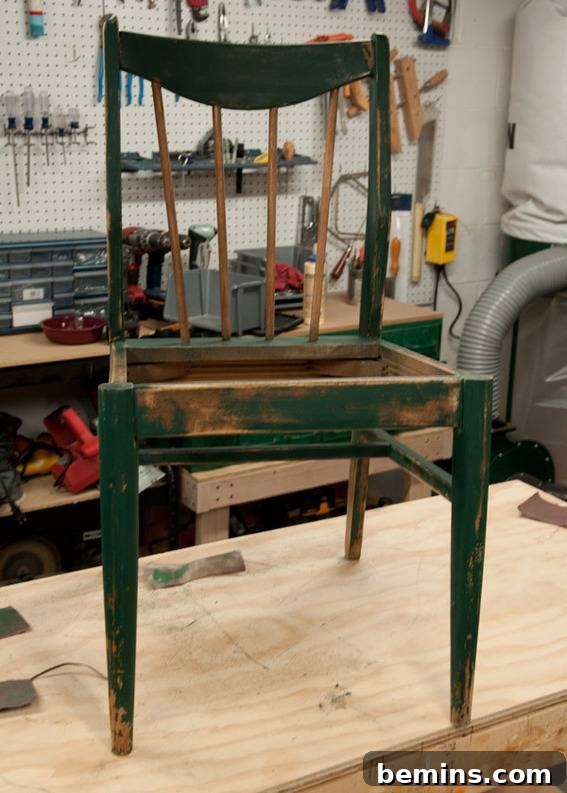 An old wooden chair that was once painted green but is now chipped and peeling sits on top of a table in a workshop. Sanded down and ready for paint.