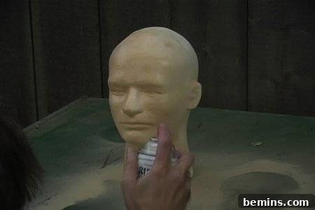 Rubber human face being molded on a metal stand by a person in a small booth.