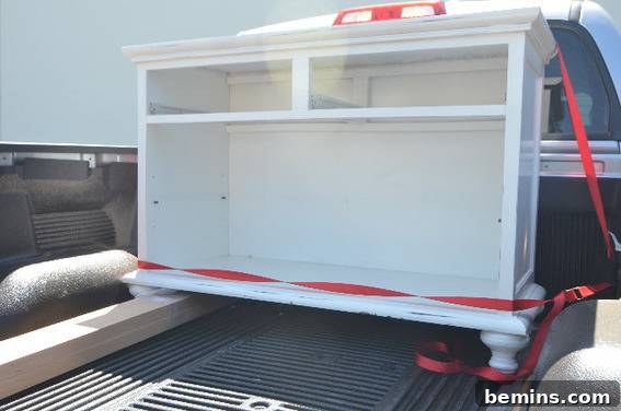 A white wooden cabinet with its drawers and shelves removed is strapped into the back of a pickup truck, showing a cabinet ready for transformation.