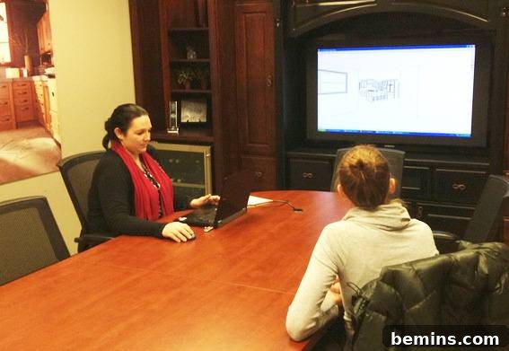 Woman sitting at a table with a computer while another woman watches a television screen.