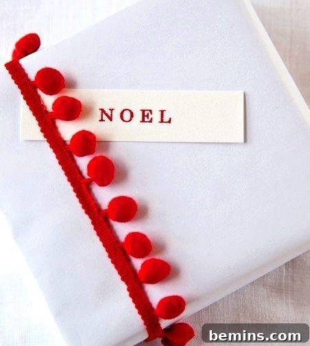 A gift covered in white cloth and a red ribbon with fuzzy red balls and a noel tag slipped under the ribbon.