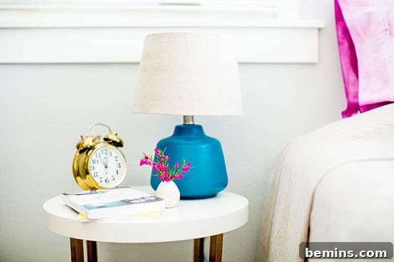 Lamp, clock and book is on the table besides bed.