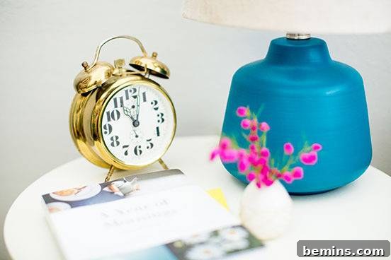 An alarm clock sits on a white table near a blue and white lamp.