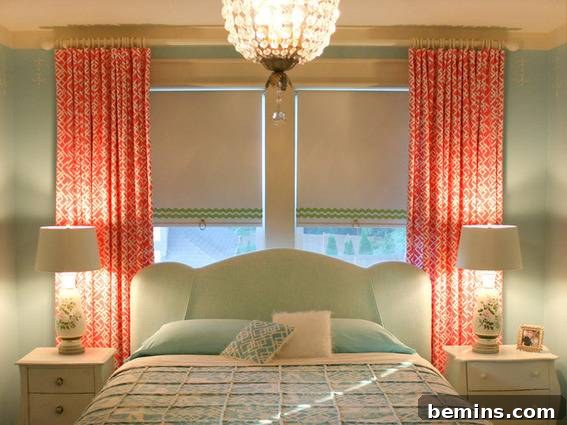 Neat bed centered between pale nightstands with lamps, in front of double window with roller shades and bright orange curtains.