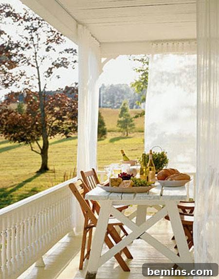 Outdoor Kitchen Dreams: 12 DIY Spaces to Inspire 11 A white porch on a sunny day with sheer curtains and a white table with brown chairs set with bread and cheeses.