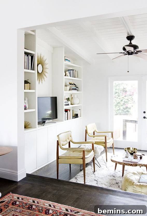 A spacious, bright white room, elegantly furnished with extensive built-in bookcases, comfortable chairs, and a sleek ceiling fan, creating an inviting and organized ambiance.