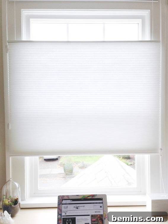 A laptop or tablet sts on a desk under a window with a white shade sitting in the center of the window.