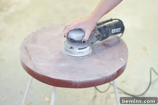 A hand using a round electric sander on the top of a round wooden stool.