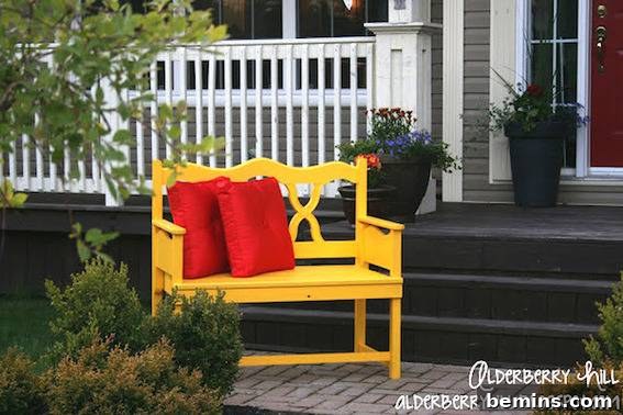Two red throw pillows sit on top of a yellow bench in front of a white fence.