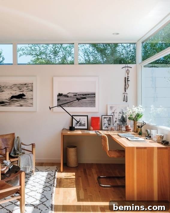A sunlit room with floor-to-ceiling rectangular windows, a minimalist wooden corner desk, and striking black and white photographs on the wall.