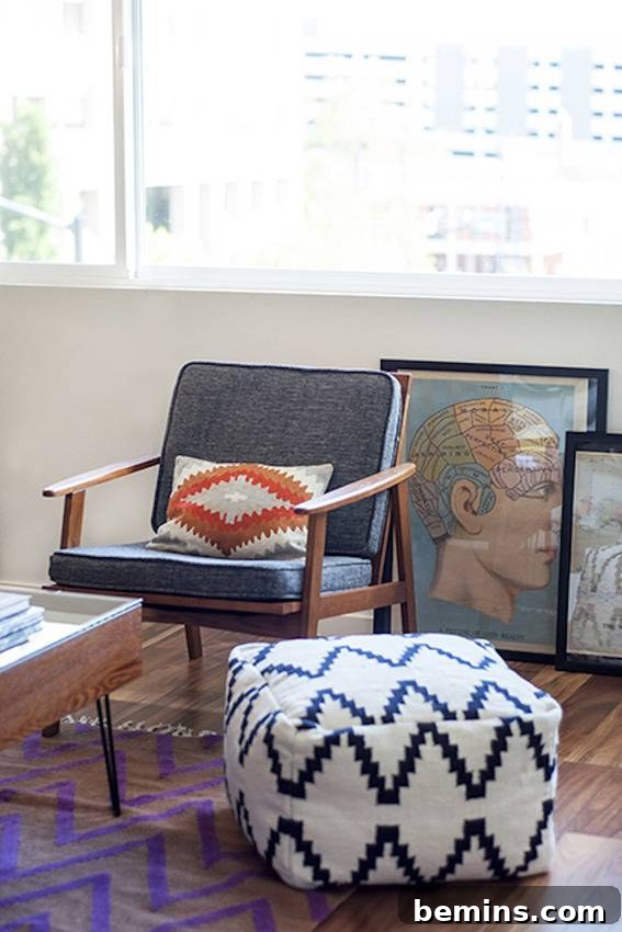 A stylish living room corner featuring a white ottoman with black zigzags, a wooden chair with grey cushions, and a tan pillow with an orange diamond pattern, next to a piece of art with a man's head on a blue background.