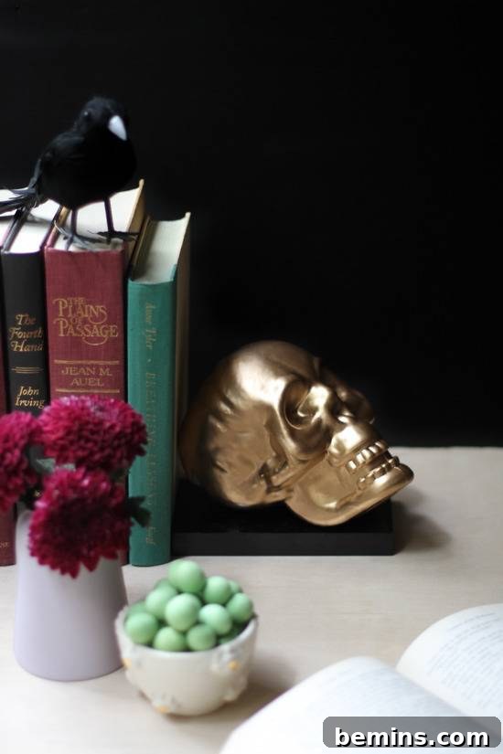 A gold skull bookend secures a red, green, and black book on a shelf, next to green mints, and red flowers.