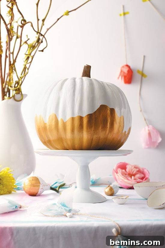 A pumpkin on a cake plate partially painted white, a white vase with yellow branches and pink and orange roses hanging upside down on the wall. An elegant no-carve pumpkin centerpiece.